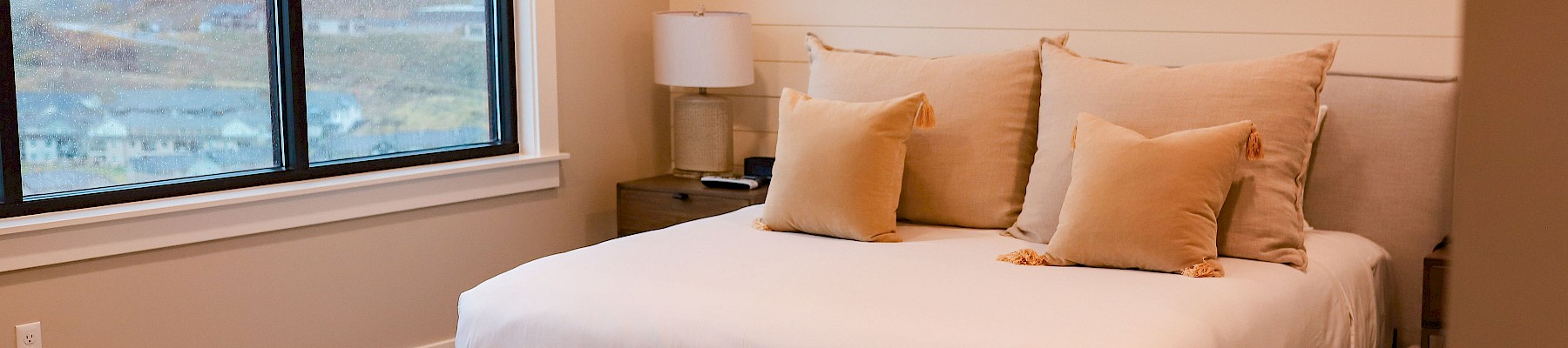 A cozy bedroom with a large bed, beige pillows, a tufted bench, nightstand with a lamp, and a window with a scenic view.