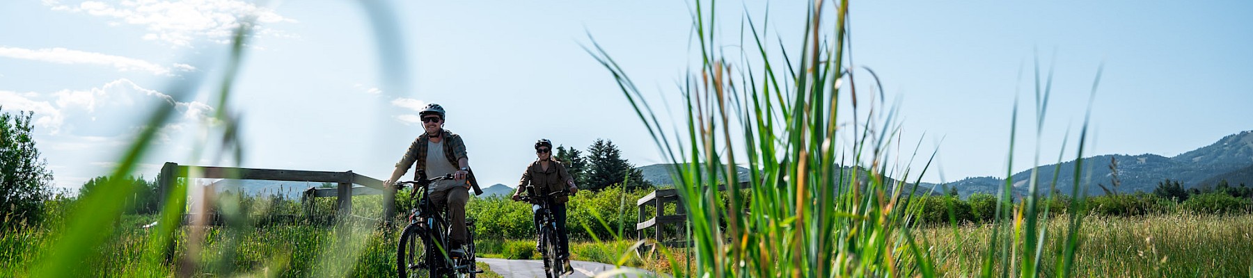 Two people are biking on a paved path amidst tall grass with a clear blue sky and distant hills in the background.