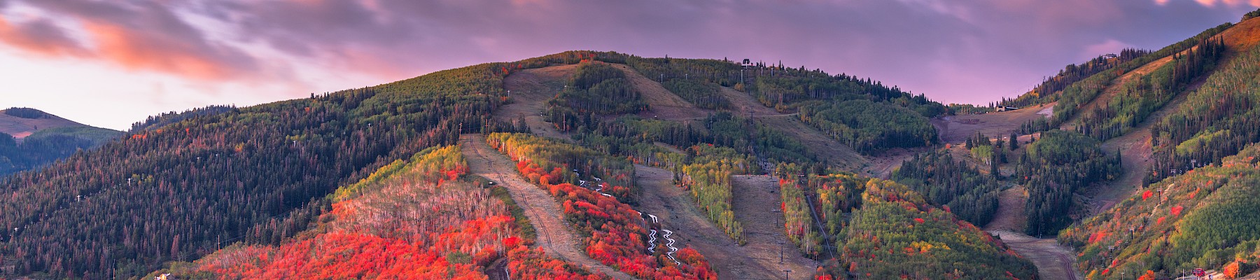 A scenic view of a town nestled in a valley with colorful autumn trees on hillsides and a dramatic cloudy sky at sunset.