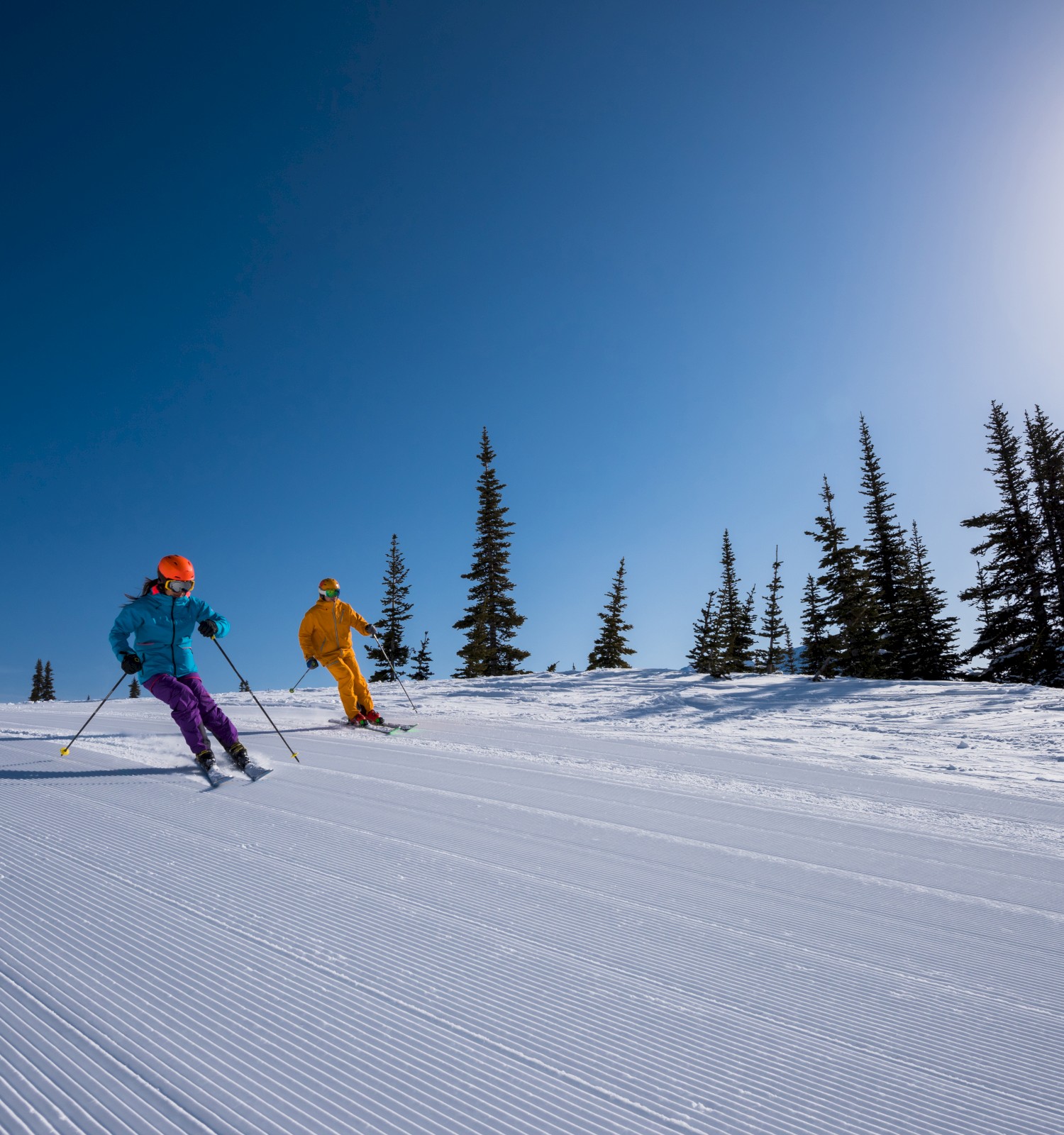 Two children in colorful ski gear are skiing down a groomed snowy slope under a bright blue sky with pine trees in the background.