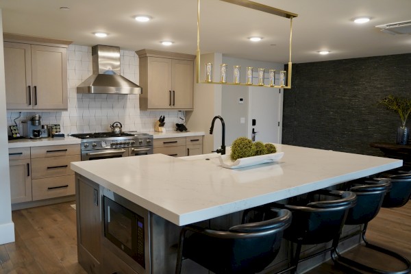 This is a modern kitchen with a large island, beige cabinets, black bar stools, and a textured dark wall.