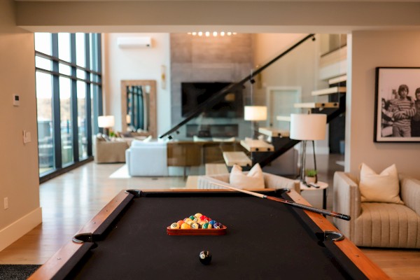 A pool table is in a modern, open living room with large windows, sofas, lamps, and contemporary decor.