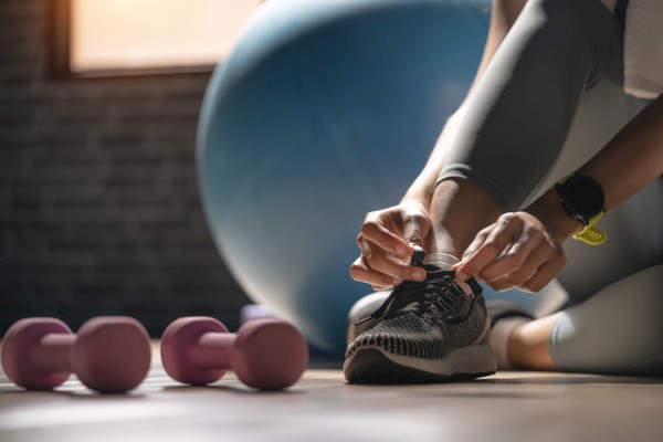 A person tying their running shoes next to pink dumbbells and an exercise ball in a gym setting.