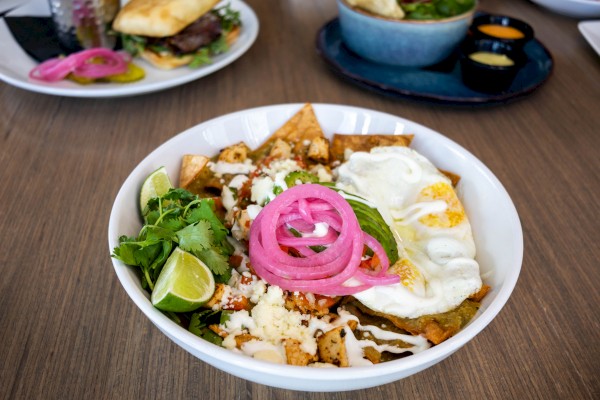 A bowl of Mexican food with fried eggs, crumbled cheese, pickled pink onions, cilantro, and tortilla chips on a wooden table.