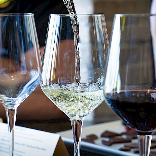 Wine glasses, one with white wine being poured, set on a table with chocolates and a menu.
