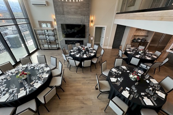 The image shows a beautifully set dining area with round tables, black tablecloths, elegant glassware, silverware, and floral centerpieces.