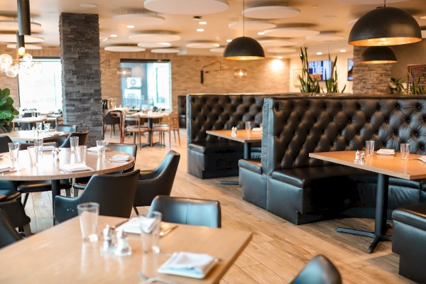 The image shows an empty modern restaurant with wooden tables, black chairs, leather booth seating, and warm lighting.