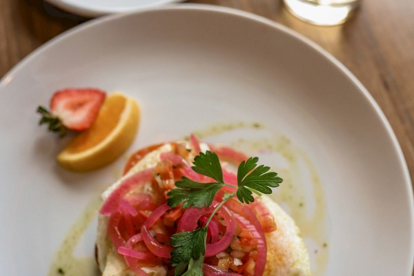 A plate featuring a dish topped with cherry tomatoes, pickled onions, and herbs, with a lemon wedge and a drink in the background.