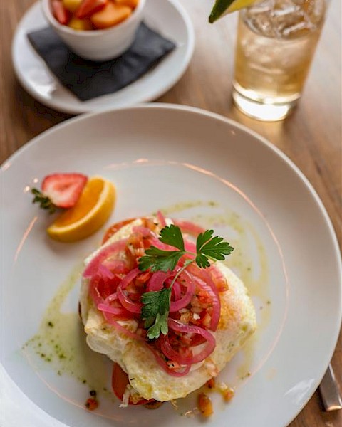 A plate with a baked dish topped with pickled onions and herbs, a lemon wedge, a small bowl of fruit salad, and a glass of iced drink with lime.