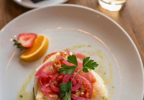A plate with a baked dish topped with pickled onions and herbs, a lemon wedge, a small bowl of fruit salad, and a glass of iced drink with lime.