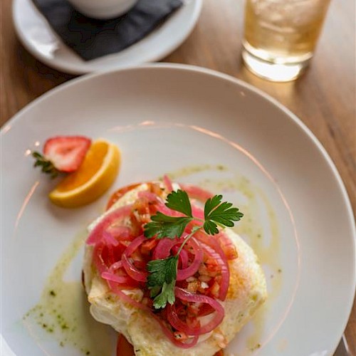 A plate with a baked dish topped with pickled onions and herbs, a lemon wedge, a small bowl of fruit salad, and a glass of iced drink with lime.