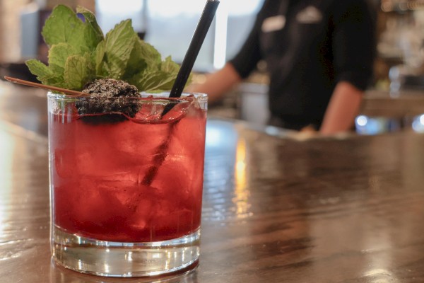A vibrant red cocktail garnished with mint and blackberries on a wooden bar counter in a cozy cafe.