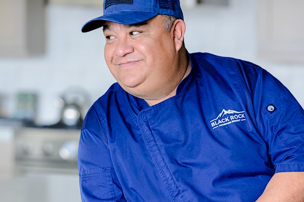 A chef in a blue uniform and cap, leaning on a kitchen counter, with a 
