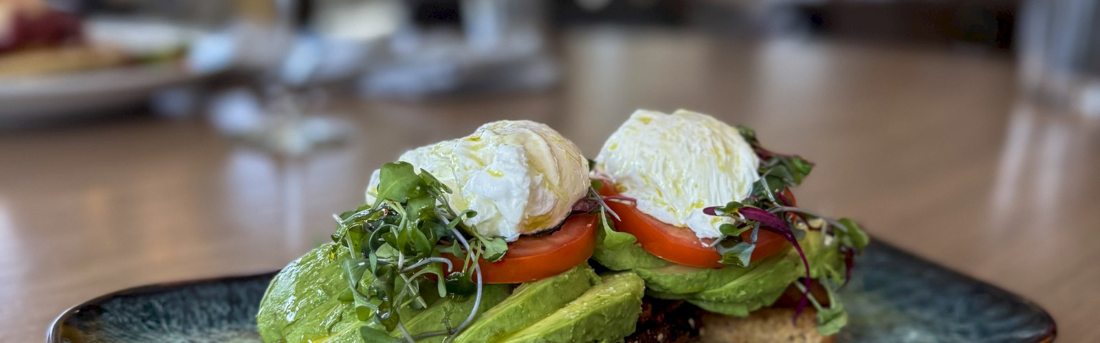 A toasted bread with avocado, tomato, microgreens, poached eggs, and bacon served on a black plate.