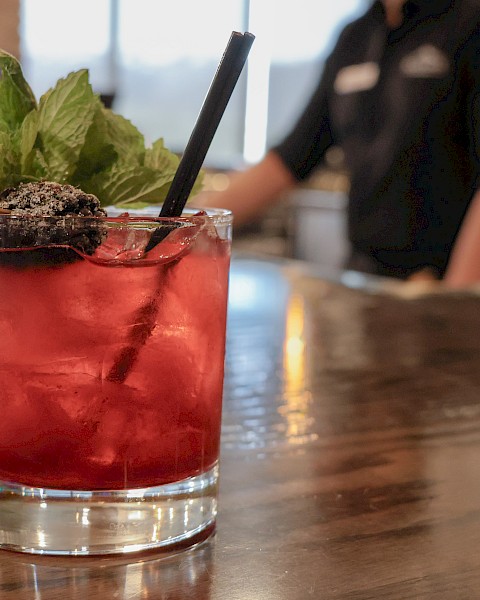 A close-up of a red cocktail garnished with mint and blackberry, placed on a wooden bar counter with a blurred bartender in the background.