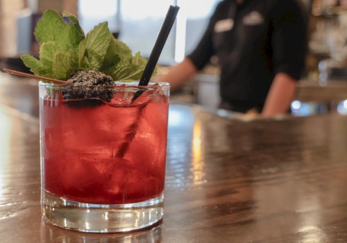 A close-up of a red cocktail garnished with mint and blackberry, placed on a wooden bar counter with a blurred bartender in the background.