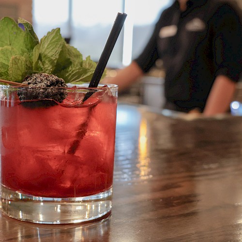 A close-up of a red cocktail garnished with mint and blackberry, placed on a wooden bar counter with a blurred bartender in the background.