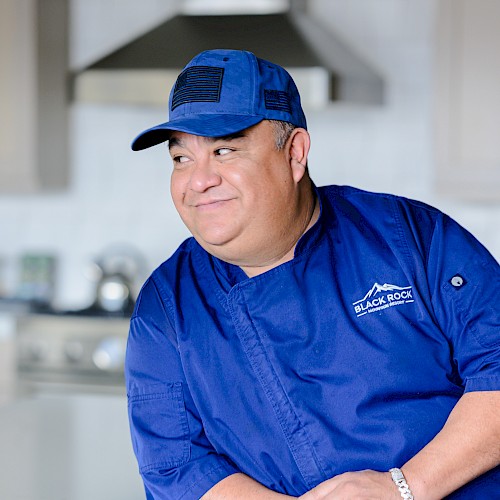 A man in a blue chef's uniform and cap, leaning on a kitchen counter, with a 