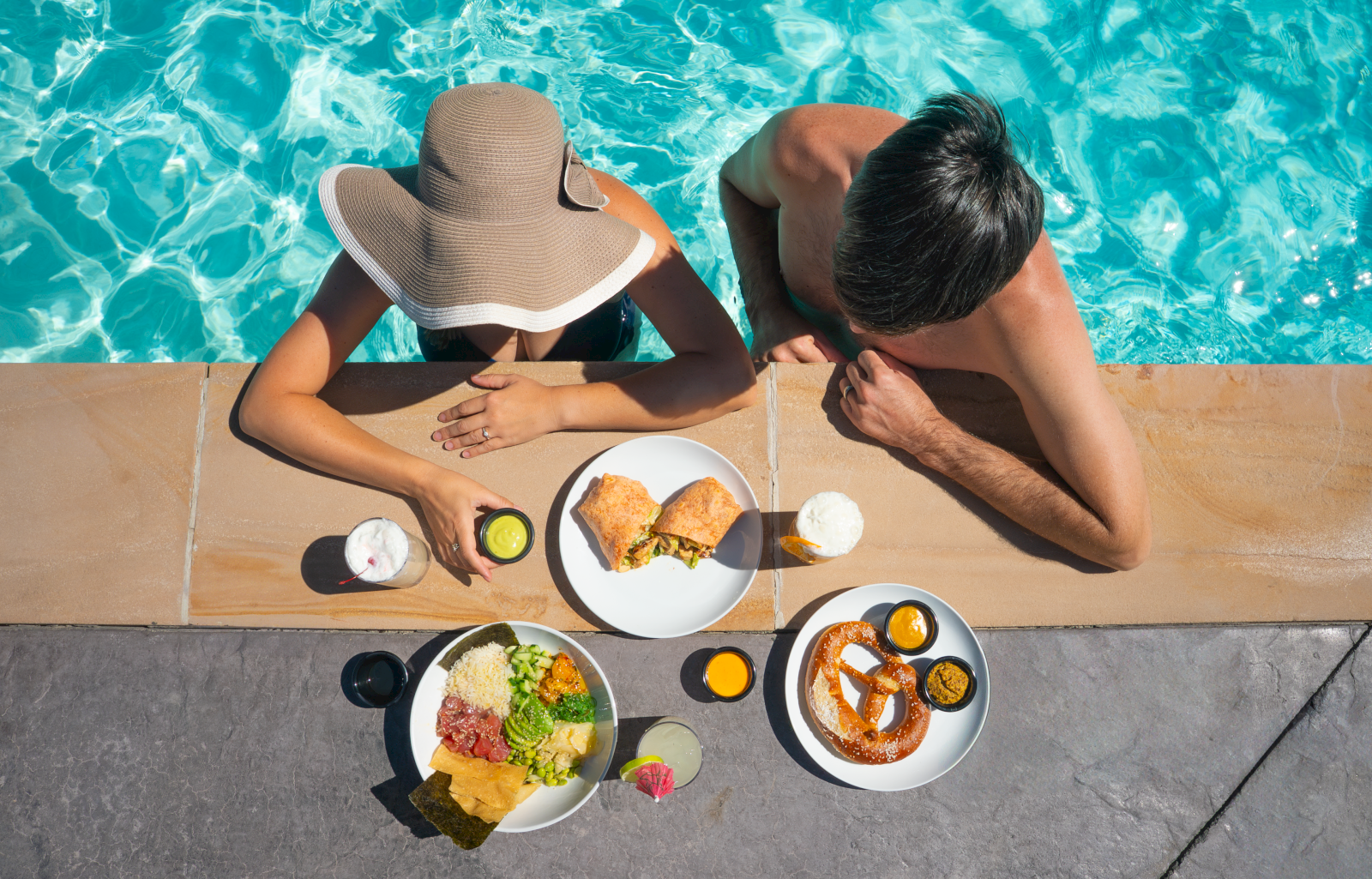 A couple enjoys food and drinks poolside, with the woman wearing a sun hat and the man leaning on the pool's edge in the image.