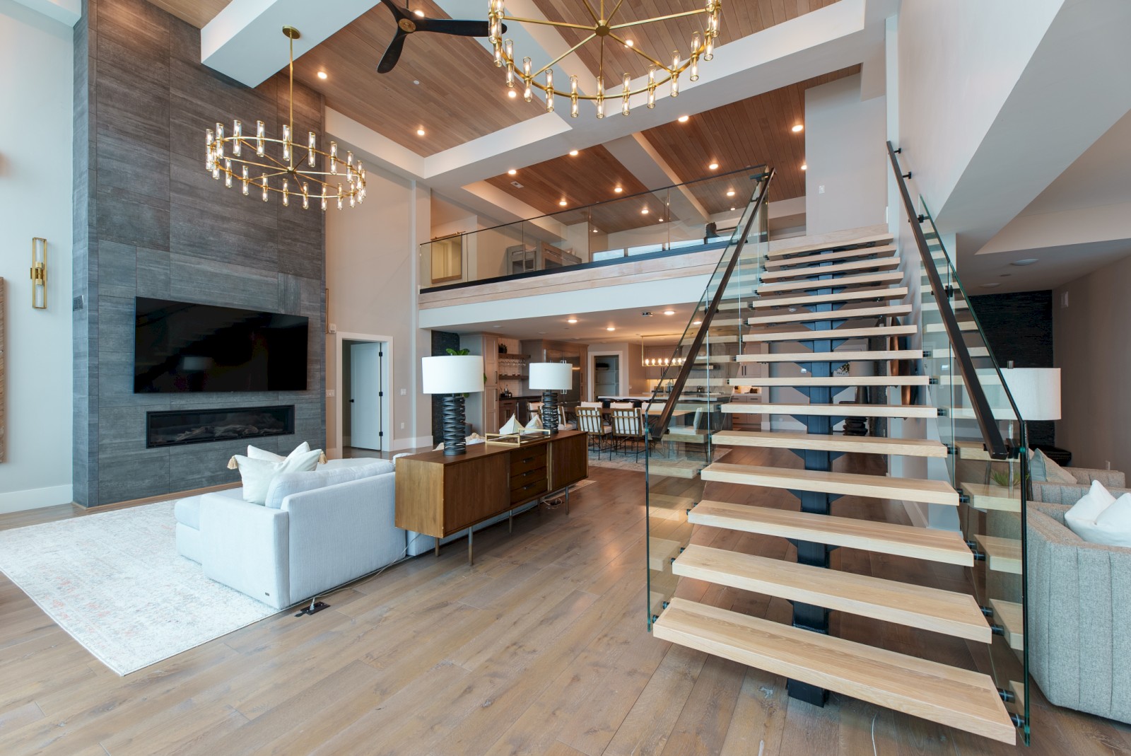 This image shows a modern, spacious living room with a high ceiling, staircase, large TV, and contemporary furniture.