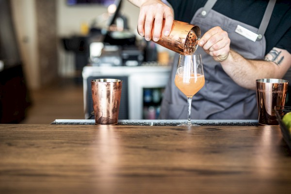 A bartender is pouring a pink cocktail into a glass, with copper-colored cups on the bar in this professional bar setting.
