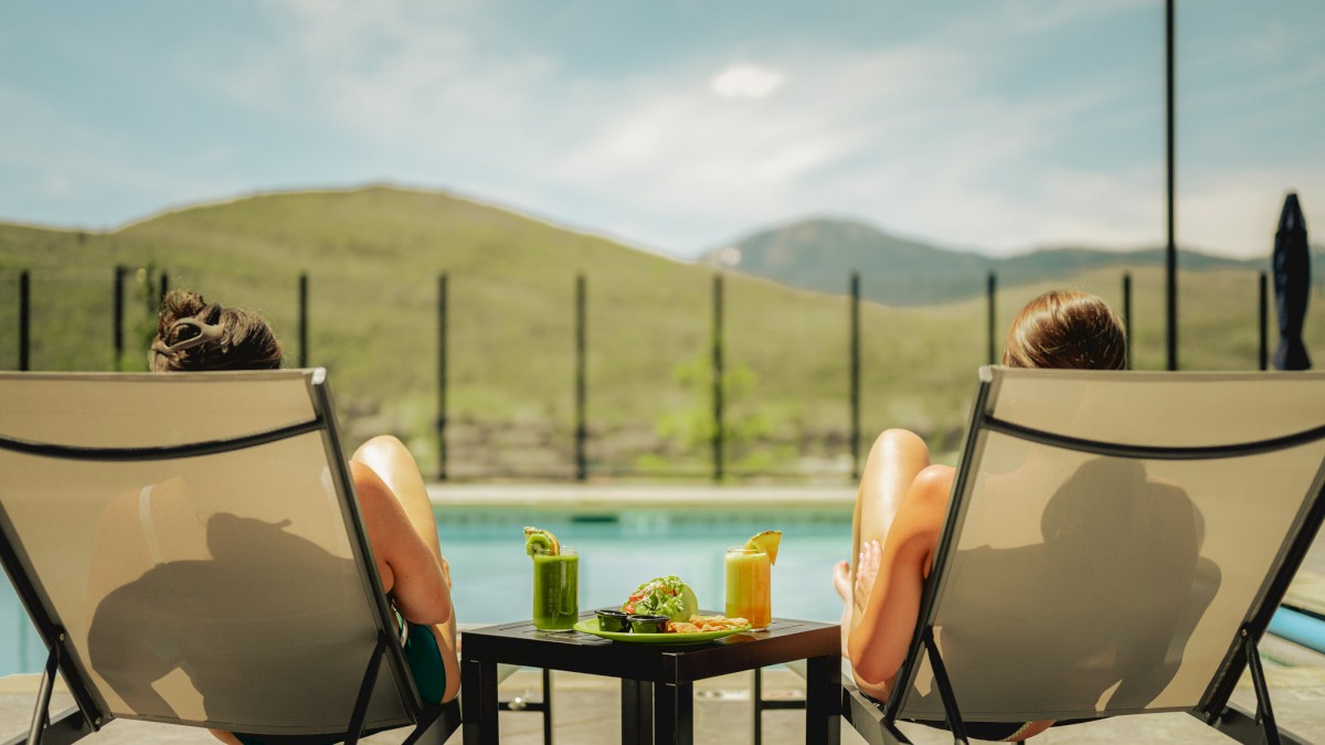 Two women relax by a pool, enjoying drinks with snacks, overlooking a scenic green mountain landscape.