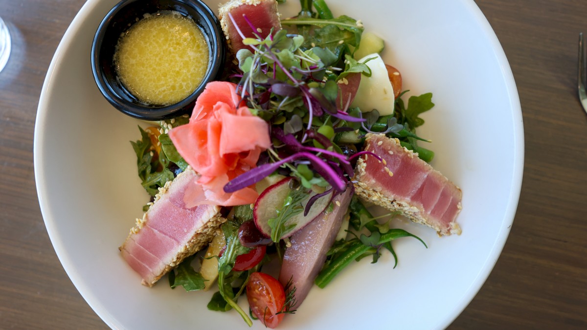 A plate with mixed greens, seared tuna, sliced vegetables, pickled ginger, and a small bowl of dressing or sauce.