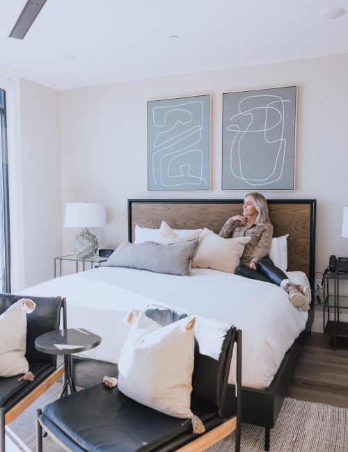 A woman relaxes on a neatly made bed in a modern, stylish bedroom with abstract art and minimal decor.