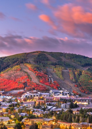 A town with vibrant autumn-colored trees and surrounding mountains under a pink-tinged sky at sunset.