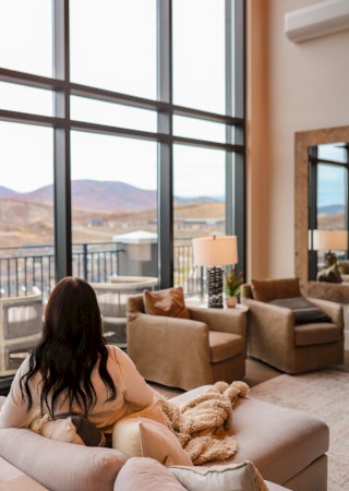 A person sits on a couch, overlooking mountains through large windows in a cozy living room with modern furnishings.