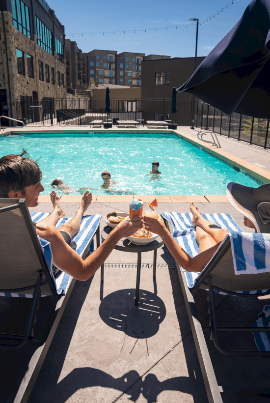People relaxing by a pool, enjoying drinks and snacks under sunny skies.