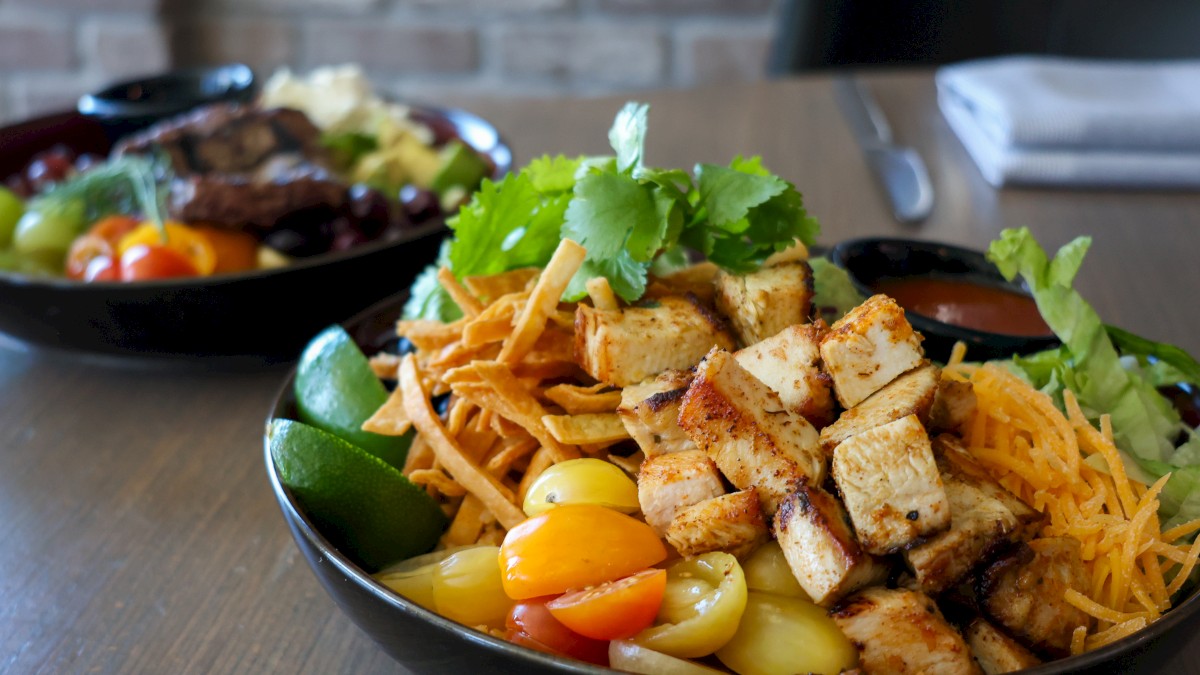 Two bowls of colorful, fresh Mexican-style salads with grilled chicken, shredded cheese, vegetables, and lime slices on a wooden table.