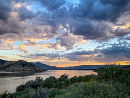 A scenic landscape with a river, green shrubs in the foreground, and a dramatic sky with clouds during sunset or sunrise.