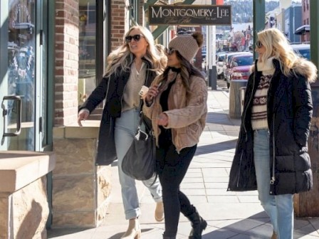 Three women are walking and talking on a city sidewalk, dressed warmly with sunglasses and cozy jackets on a sunny day.
