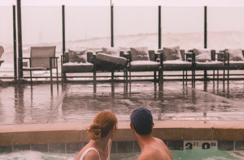A couple is sitting in a hot tub by a window with chairs outside, overlooking a snowy landscape in a cozy indoor setting.