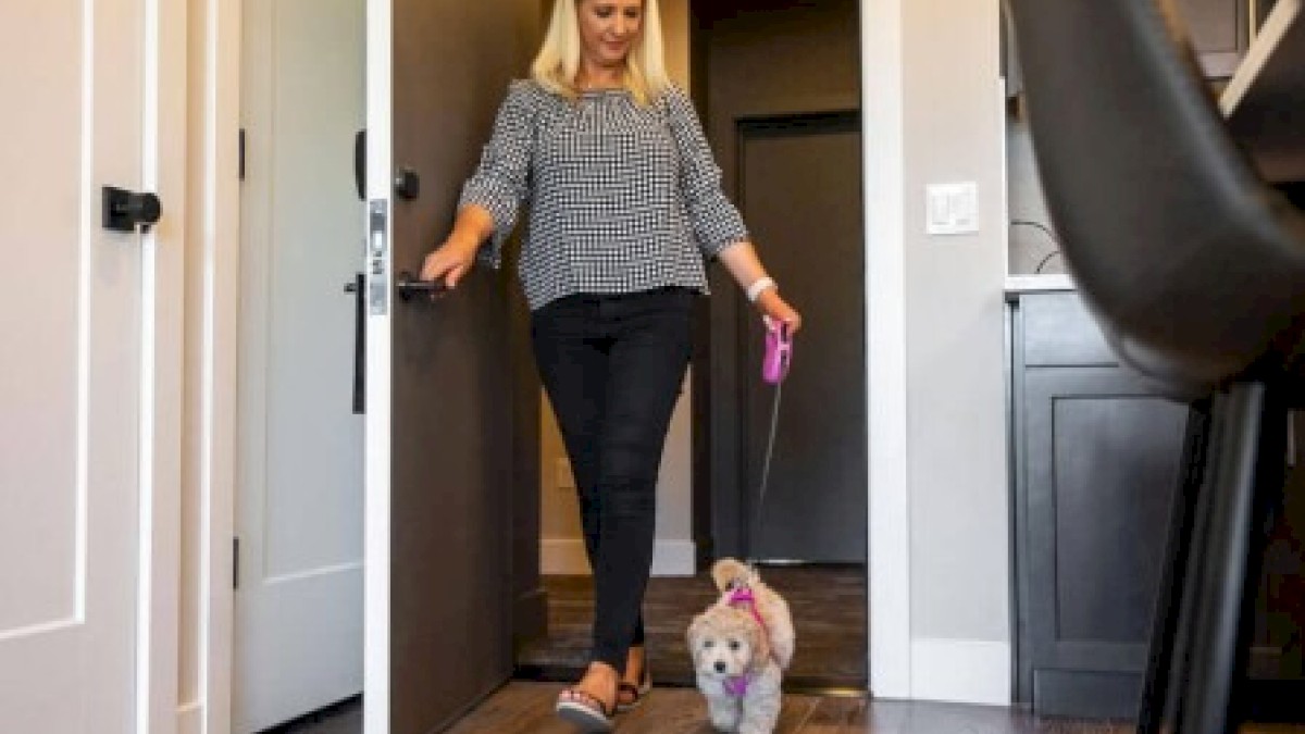 A woman walks into a room with a small, fluffy dog on a leash in a modern home interior.