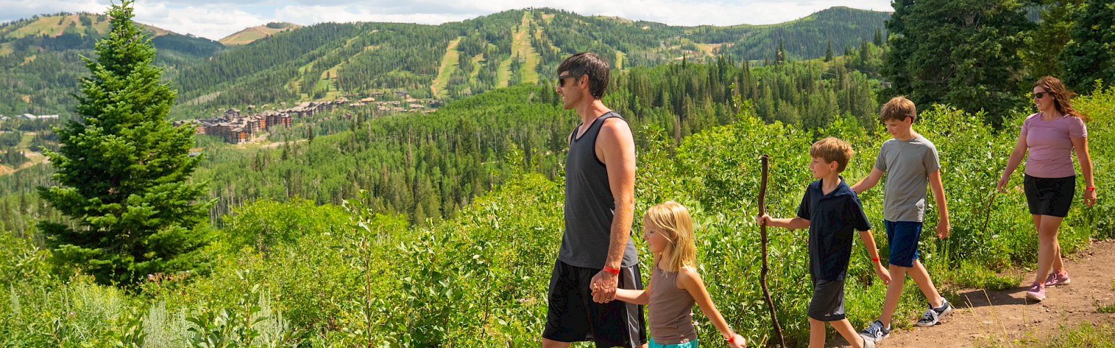 A family hikes through a lush green trail in a scenic mountainous area during a sunny day.