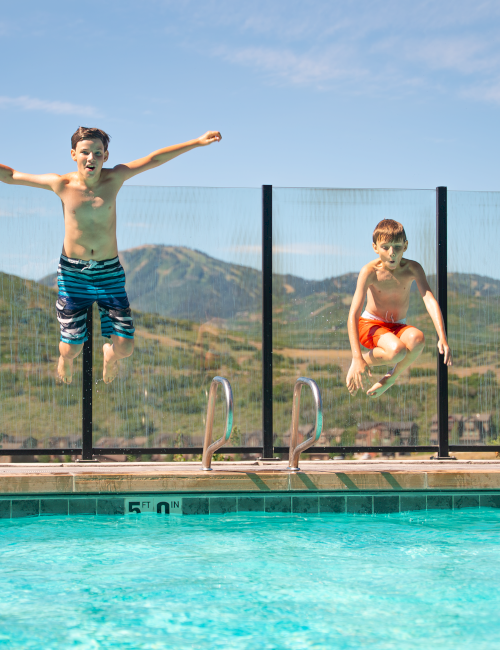 Two boys are jumping into a pool from the deck, wearing swim trunks, with mountains and blue sky in the background.