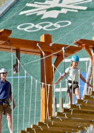 People are walking on a wooden suspension bridge at a ski resort, with a ski jump and Olympic logo visible in the background.