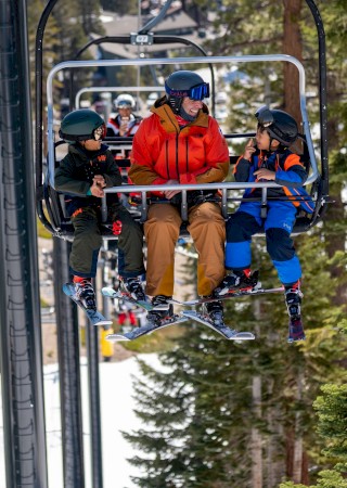 A group of three people, including two children and an adult, are riding a ski lift with a snowy mountain and pine trees in the background.