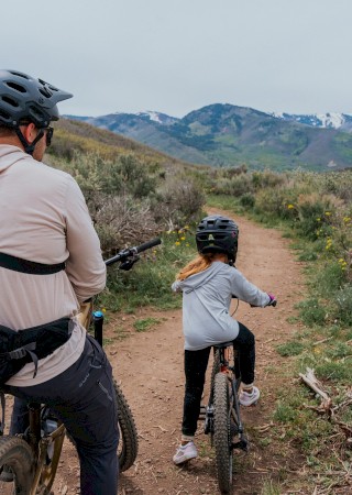 A family with helmets rides mountain bikes on a dirt trail through a scenic, hilly landscape with mountains in the background.