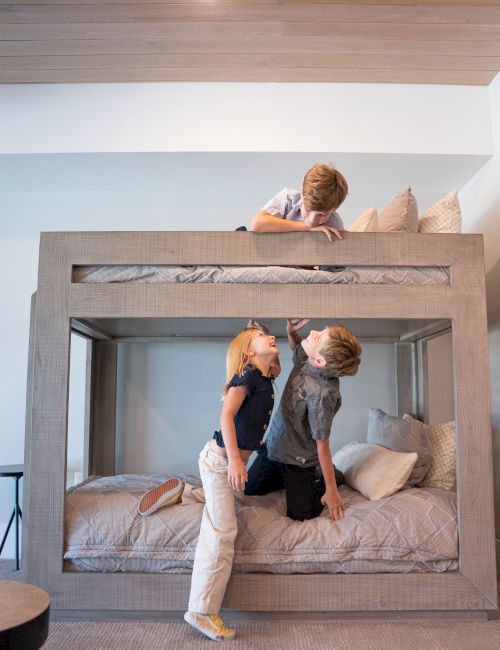 Three children play inside a bed, pretending it's a house, with one looking from above and two interacting on the mattress below.