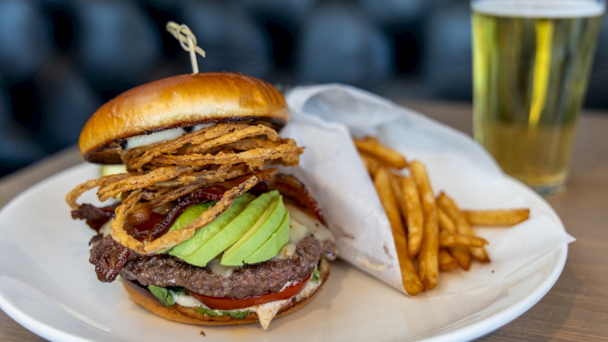 A burger with fried onion rings, avocado, tomato, lettuce, bacon, cheese, and pickles, served with fries and a glass of beer.