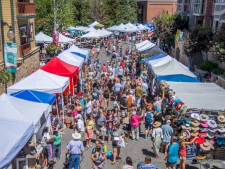 A busy outdoor street market with vendor tents, shoppers, and various goods, in a lively urban setting on a sunny day.