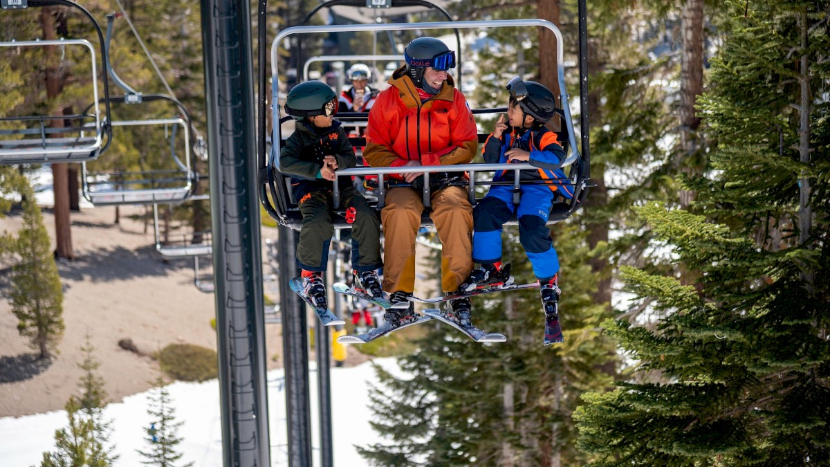 People are enjoying a ski lift ride together in a snowy forested area, with two children and an adult all wearing helmets and winter gear.