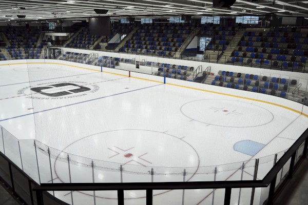 An empty indoor hockey rink with blue and black seating in the stands and a protective net in front of the ice.