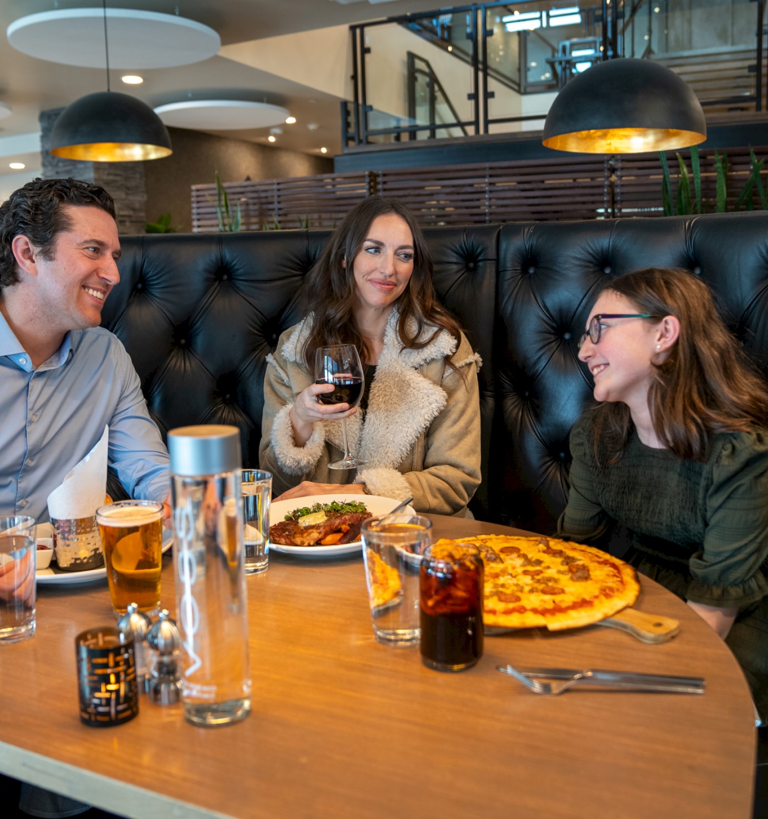 Three people are enjoying a meal and drinks at a restaurant booth, sharing laughter and good conversation.