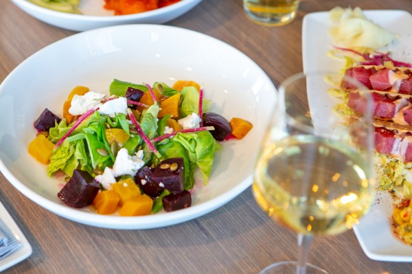 A colorful mixed salad with greens, beets, yellow peppers, cheese, and seeds, served with a glass of white wine.