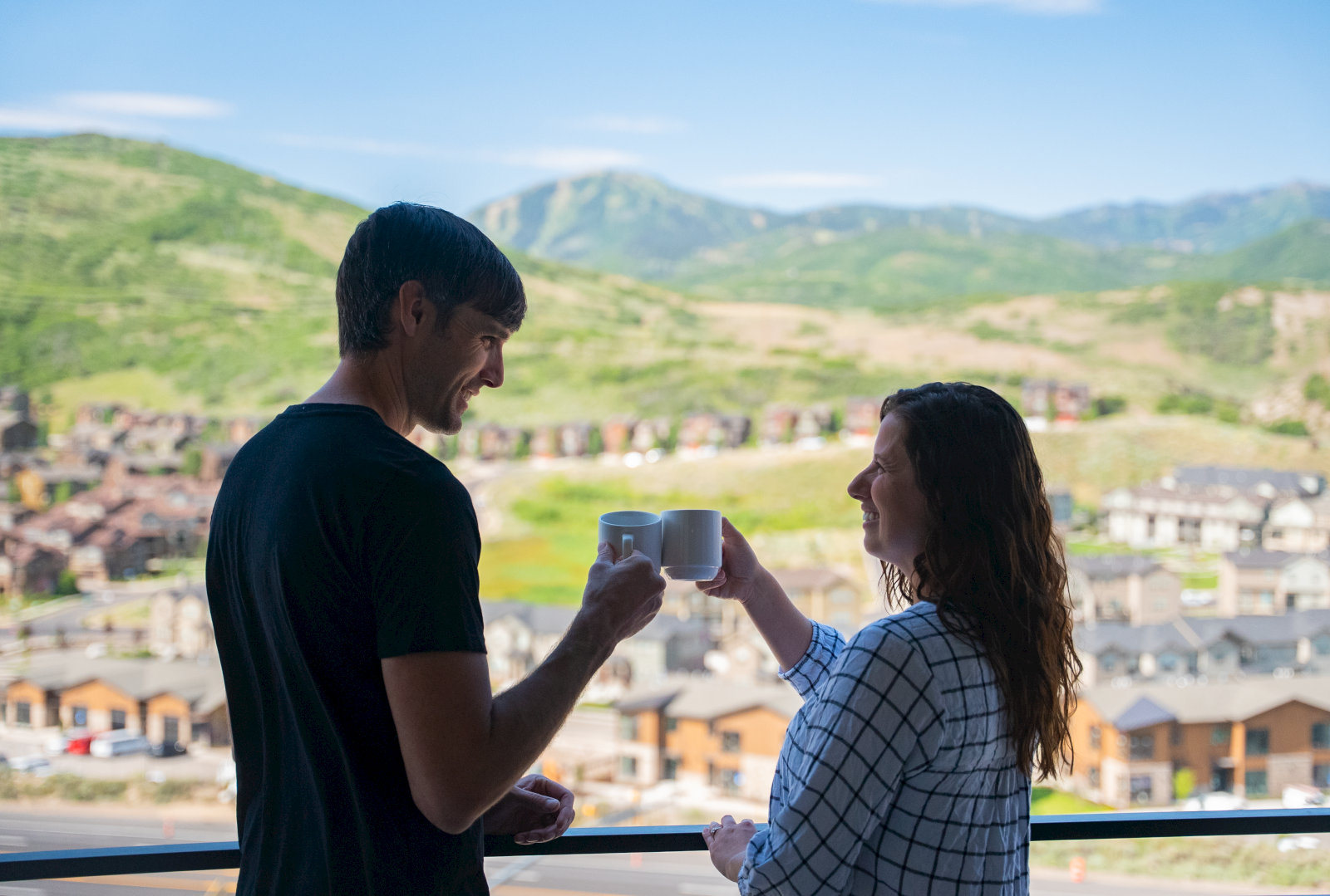 A man and woman toast with coffee mugs on a balcony, with mountains and a small town in the background during daytime.