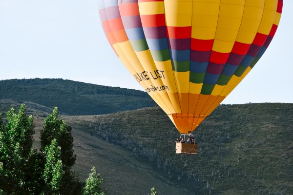 A hot air balloon with colorful stripes floats over a green mountainous landscape under a clear sky.
